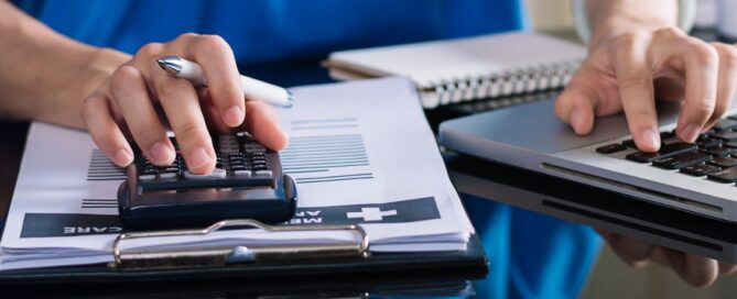 Hands using a calculator and laptop on a desk with documents, possibly related to grant writing.