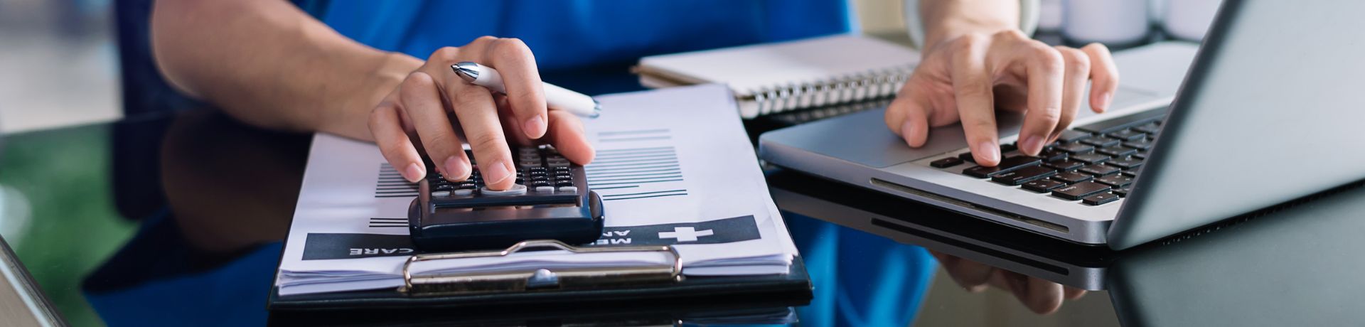 Hands using a calculator and laptop on a desk with documents, possibly related to grant writing.