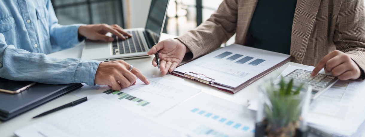 Colleagues discussing charts and graphs on a desk, focusing on grant writing. A laptop, clipboard, and calculator are in use.