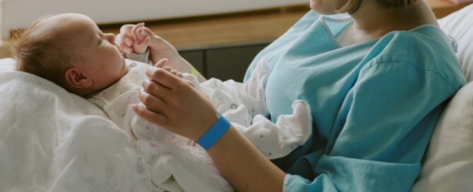 Mother in hospital gown cradles newborn wrapped in a white blanket, sitting on a hospital bed, sunlight streaming through a window.