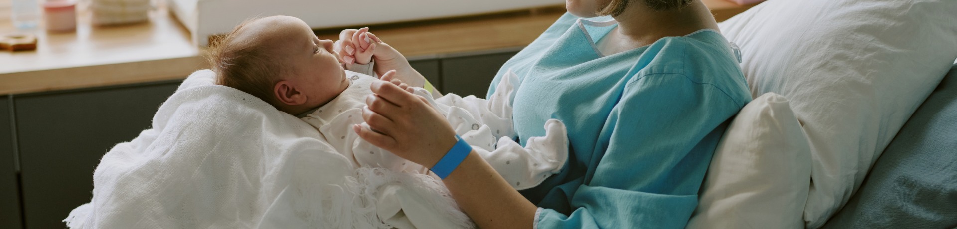 Mother in hospital gown cradles newborn wrapped in a white blanket, sitting on a hospital bed, sunlight streaming through a window.