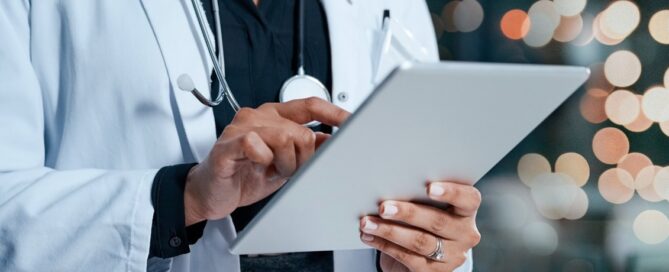 Doctor using a tablet, possibly for grant writing, against a blurred background of city lights.