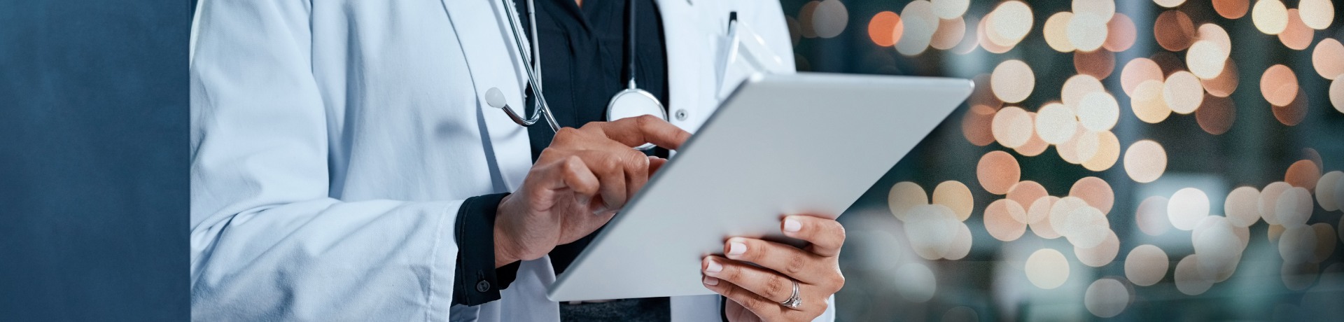 Doctor using a tablet, possibly for grant writing, against a blurred background of city lights.