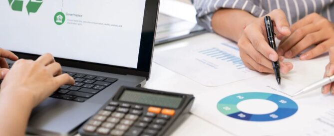 Hands typing on a laptop with data charts and a calculator nearby, suggesting teamwork in grant writing and financial planning.